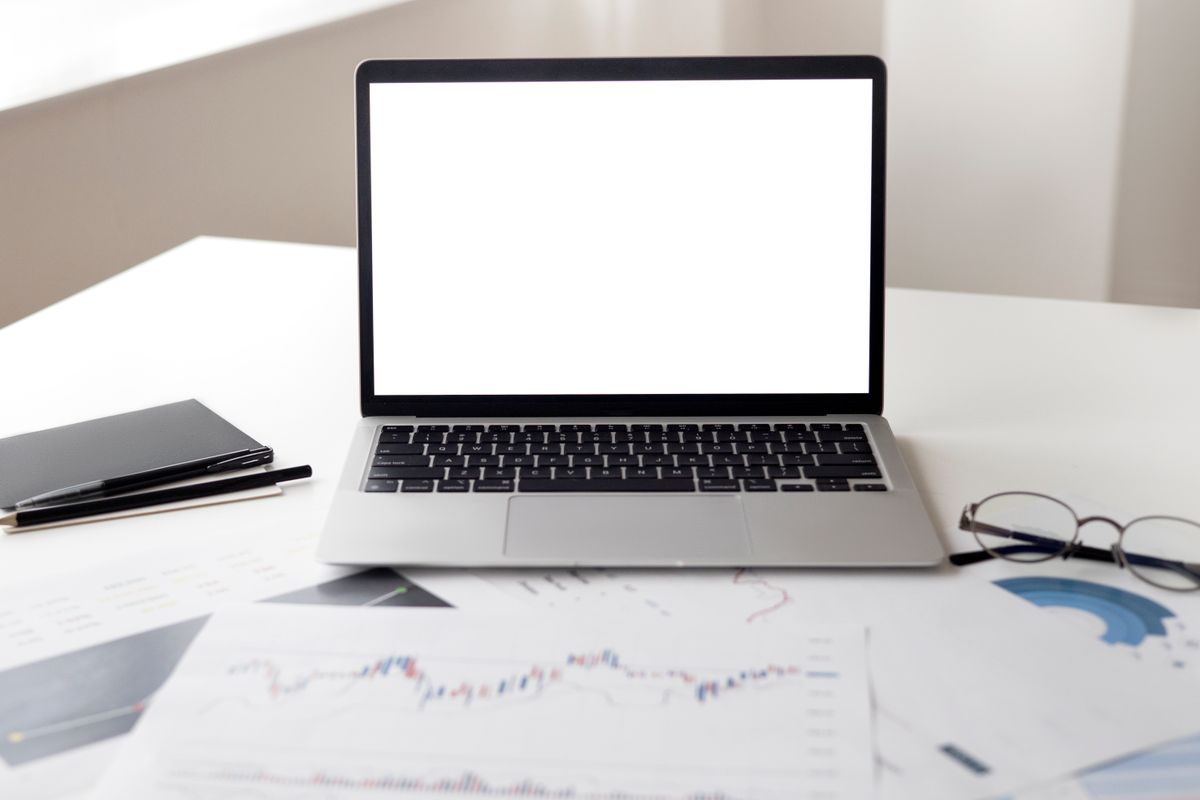 A laptop beside notebooks, documents and glasses on a desk.